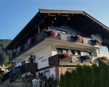 A picturesque house in alpine style with flowering balcony boxes. Green mountains are visible in the background.
