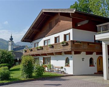 Ein traditionelles alpines Haus mit Holzbalustrade und Blumenkästen. Im Hintergrund sind grüne Hügel und ein Kirchturm zu sehen.
