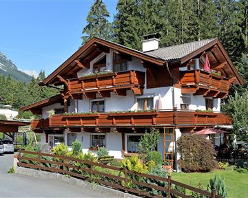 Ein charmantes Holzhaus im alpinen Stil, umgeben von Bäumen und schöner Vegetation. Die Balkone sind mit Blumen dekoriert und das Gebäude strahlt eine einladende Atmosphäre aus.