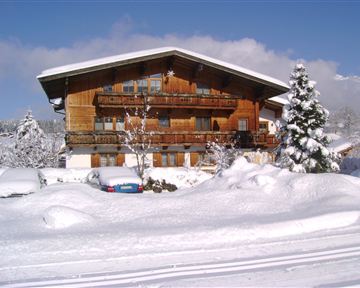 Ein Holzhaus im Winter, umgeben von viel Schnee. Strahlend blauer Himmel und schneebedeckte Bäume sorgen für eine idyllische Atmosphäre.