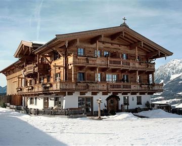 Ein wunderschönes alpenländisches Holzhaus, umgeben von Schnee und Bergen. Die Architektur ist traditionell mit Balkonen und Holzverzierungen.