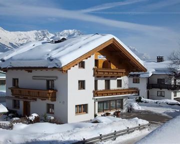 Ein charmantes Haus im alpinen Stil, umgeben von Schnee. Im Hintergrund sind schneebedeckte Berge und ein blauer Himmel zu sehen.
