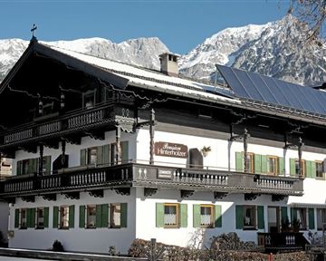 A traditional alpine house with a beautiful balcony and green shutters. Snow-covered mountains are visible in the background.