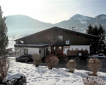 A charming house in the snow, surrounded by mountains and trees. The sun is shining, creating a warm atmosphere.