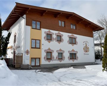 Ein traditionelles Haus mit einem Holzgeschoss und bunten Fenstern. Der Schnee liegt vor dem Gebäude, das von einem klaren Himmel umgeben ist.