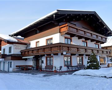 Ein traditionelles Haus im alpenländischen Stil, umgeben von Schnee. Die Fassade ist mit Holzverzierungen und einem Balkon gestaltet.
