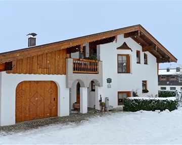 Ein charmantes Haus im alpinen Stil, umgeben von Schnee. Der Dachstuhl ist aus Holz, und es gibt eine kleine Terrasse mit Pflanzen.