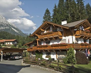 A traditional chalet surrounded by trees and mountains. The balconies are adorned with flowers, and the surroundings are peaceful and picturesque.