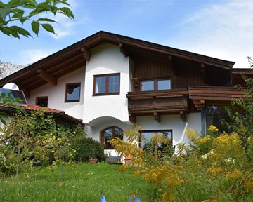 A charming house with wood siding and large windows in a green garden. Gentle hills are visible in the background.