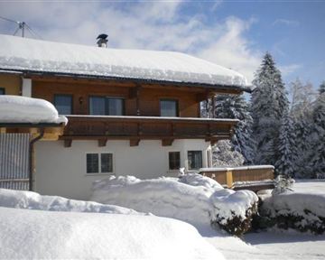 A cozy house in the snow with a wooden balcony. All around, the landscape is surrounded by snow-covered trees.