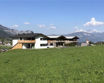 A modern building is situated in a green landscape. In the background, mountains and a small church tower are visible.