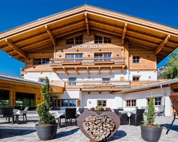 A charming wooden building in alpine style with a large porch.  
Patio furniture and plants invite you to linger.