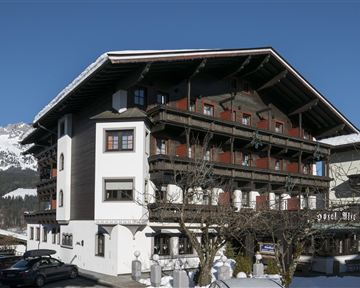 A traditional Alpine hotel with multiple balconies and a large roof. Snow lies around the building, and the mountains are visible in the background.