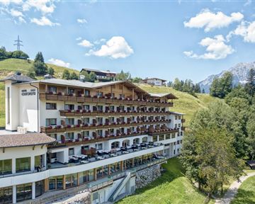 A modern hotel with several balconies, surrounded by green hills and mountains. The sky is clear and sunny.