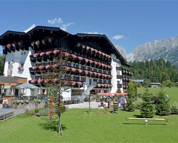 A picturesque hotel building with balconies adorned with flowers. In the background, green meadows and impressive mountains can be seen.