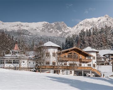 A charming house in a snowy landscape, surrounded by mountains. The sky is clear and there are many trees nearby.