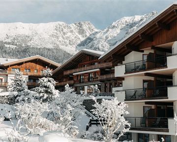 Eine malerische Winterlandschaft mit schneebedeckten Hütten und hohen Bergen im Hintergrund. Die Häuser haben holzverkleidete Fassaden und sind von schneebedeckten Bäumen umgeben.