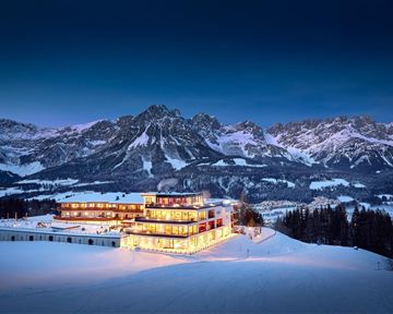 Ein modernes Hotel in einer schneebedeckten Landschaft mit beeindruckenden Bergen im Hintergrund. Die Fassade des Hotels leuchtet warm im Dämmerlicht.