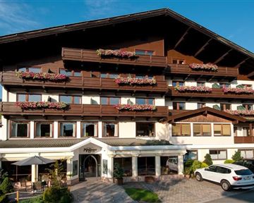 Ein charmantes Hotelgebäude mit Balkonen und Blumenschmuck. Die sonnige Umgebung und die Berglandschaft verleihen dem Ort eine einladende Atmosphäre.
