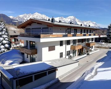 A modern building in the snow with balconies and large windows. In the background, impressive mountains and blue sky can be seen.