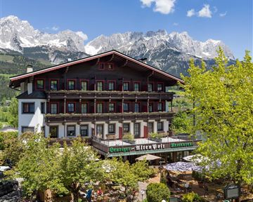 Ein charmantes Hotel mit traditioneller Architektur, umgeben von grünen Bäumen. Im Hintergrund erheben sich majestätische Berge unter einem klaren Himmel.