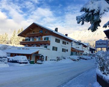 Eine verschneite Straße mit mehreren Häusern in einem Winterdorf. Die Umgebung ist malerisch mit Schnee bedeckten Bäumen und einem klaren Himmel.
