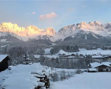 Eine winterliche Landschaft mit verschneiten Bergen im Hintergrund. Kleine Häuser sind in der Nähe sichtbar, während die Sonne die Bergspitzen beleuchtet.