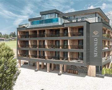 A modern hotel building with wooden elements and large windows. In the background, mountains and a green area are visible.