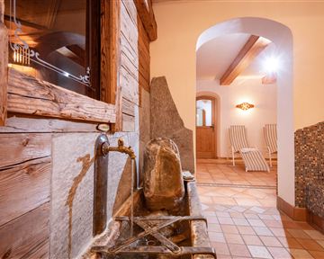 Ein gemütlicher Wellnessbereich mit einem traditionellen Brunnen aus Holz und Stein. Im Hintergrund sind Liegen und eine einladende Atmosphäre sichtbar.