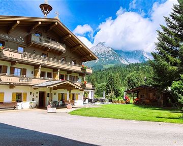Ein traditionelles Alpenhaus mit mehreren Balkonen und schönen Blumen. Im Hintergrund sind grüne Wiesen und Berge zu sehen.