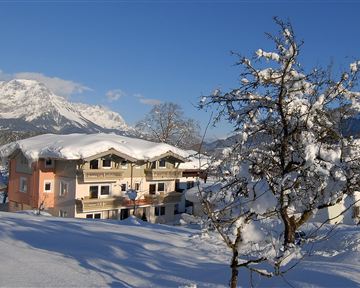 Ein gemütliches Haus im Schnee mit einem strahlend blauen Himmel. Die Umgebung ist von schneebedeckten Bergen und Bäumen geprägt.
