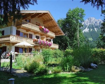 Ein schönes Holzhaus mit Balkonen, geschmückt mit Blumen. Im Hintergrund sind mit Bäumen bewachsene Berge zu sehen.