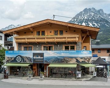 Ein gemütliches Holzgebäude mit einem großen Werbeschild. Im Hintergrund sind beeindruckende Berge und ein teilweise bewölkter Himmel zu sehen.