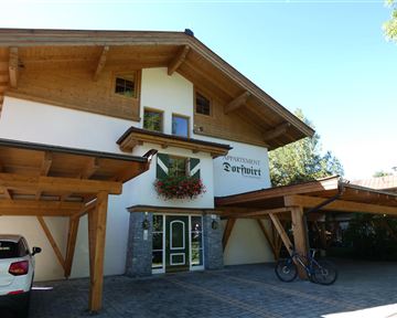 A cozy apartment building with wooden cladding and blooming plants. In the foreground, there is a car and a bicycle.