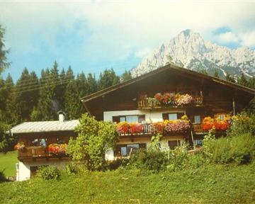 A traditional mountain hut with colorful flower balconies. In the background, high mountains rise beneath a blue sky.