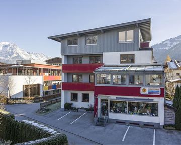 Ein modernes Gebäude mit roter und grauer Fassade, umgeben von Bergen. Es gibt einen Parkplatz und einen Eingangsbereich mit Glasverkleidung.
