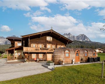 Ein traditionelles Holzhaus in den Bergen, umgeben von grünen Wiesen. Im Vordergrund steht ein ungewöhnliches, rundes Gebäude, das zur Unterkunft dient.