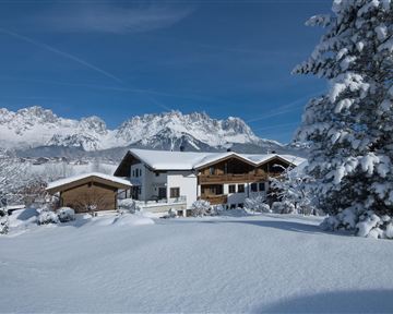 A picturesque winter landscape with snow-covered hills and mountains in the background. The wooden houses rest gently in the snow and radiate coziness.