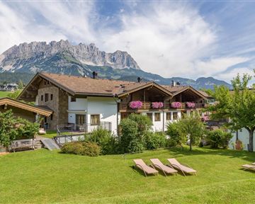 A tranquil chalet in the greenery with blooming flowers. In the background, the mountains rise majestically under a clear sky.