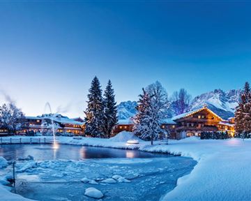 A picturesque winter landscape with snow-covered trees and a frozen pond. In the background, cozy cabins and mountains can be seen under a clear blue sky.