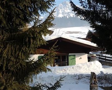 A cozy mountain cabin in the snow, surrounded by spruces and with a majestic mountain backdrop in the background. The sky is clear and the landscape looks peaceful.