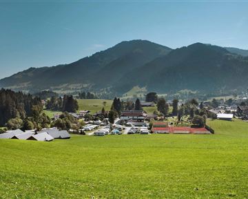 A picturesque landscape with gentle hills and a small village. In the background, mountains and a blue sky can be seen.