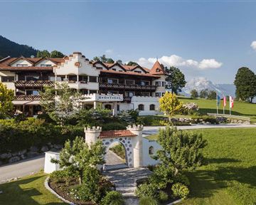 A charming hotel with traditional architecture, surrounded by green meadows and trees. In the background, mountains and a clear sky are visible.