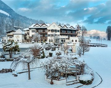 An idyllic hotel in the snow, surrounded by snow-covered trees and mountains. The sky is lightly clouded, giving the scene a calm atmosphere.