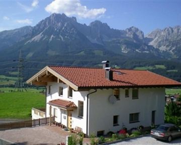 Ein schönes Haus mit rotem Dach in einer grünen Landschaft. Im Hintergrund erheben sich majestätische Berge unter einem blauen Himmel.