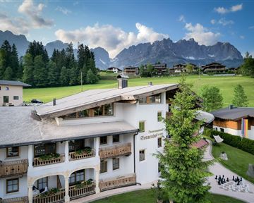 A cozy hotel in the mountains, surrounded by green meadows and tall trees. Impressive mountains can be seen in the background.