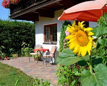 Ein gemütlicher Außenbereich mit einem Tisch und Stühlen vor einem Haus. Im Vordergrund blüht eine Sonnenblume und es gibt grünes lauschiges Gras.