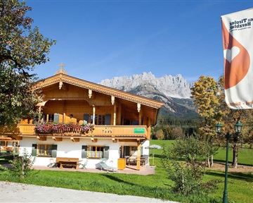 Ein traditionelles alpenländisches Holzhaus umgeben von Wiesen und Bergen. Die Fahne im Vordergrund weist auf die Region hin.