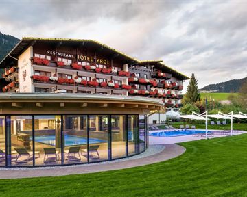 A modern hotel with a beautiful outdoor pool and well-kept garden. In the background, the mountains and a cloudy sky can be seen.