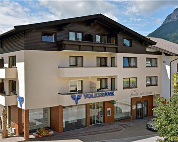 Ein modernes Gebäude mit einer Bankfiliale im Erdgeschoss. Im Hintergrund sind Berge und ein blauer Himmel zu sehen.